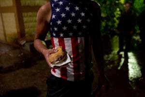 En esta imagen, tomada el 16 de octubre de 2018, un migrante hondureño vestido con una camiseta con la bandera de Estados Unidos, sostiene un sandwich en un albergue improvisado en Chiquimula, Guatemala. El presidente de Estados Unidos, Donald Trump, amenazó con cortar la ayuda a Honduras si no frena la improvisada caravana de migrantes. (AP Foto/Moisés Castillo)